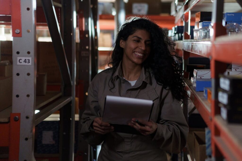Woman in jumpsuit smiling in the middle of a warehouse isle while viewing a silver tablet using Acumatica for inventory management