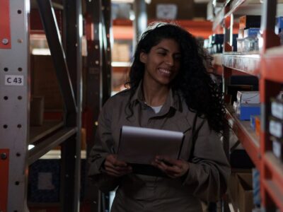 Woman in jumpsuit smiling in the middle of a warehouse isle while viewing a silver tablet using Acumatica for inventory management