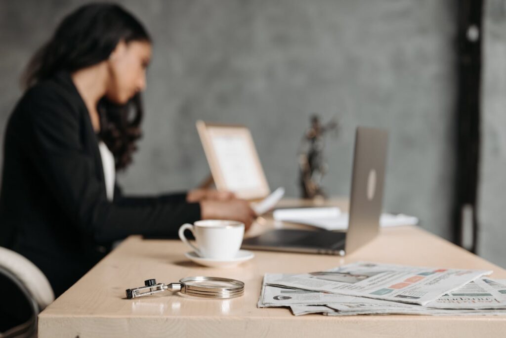 An out of focus shot of a woman in  a black suit sitting at a desk looking reports while additional reports and her laptop are in the background, as she learns to drill down in Generic Inquiries in Acumatica