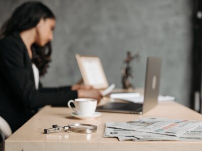 An out of focus shot of a woman in a black suit sitting at a desk looking reports while additional reports and her laptop are in the background, as she learns to drill down in Generic Inquiries in Acumatica