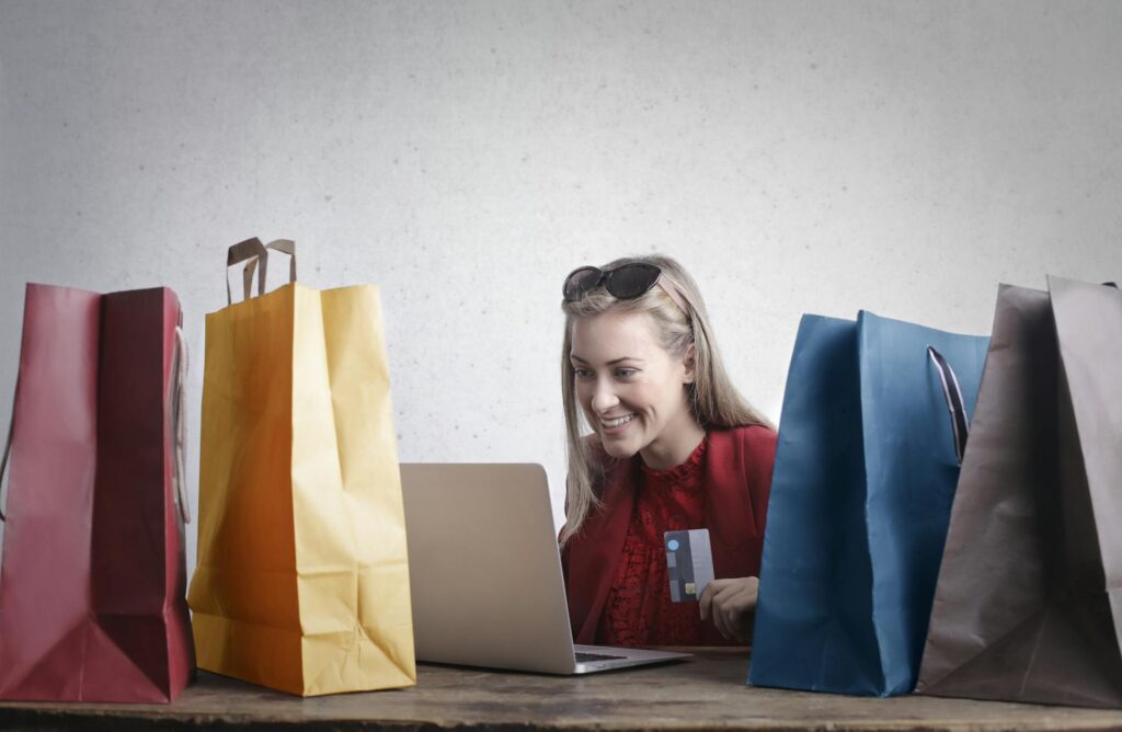 A smiling woman sitting in front of a laptop and holding  a credit card while flanked by two shopping bags each on either side, symbolizing purchases made throguh Shopify or BigCommerce integrated with Acumatica ERP for eCommerce