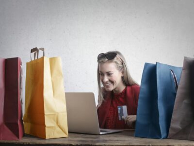 A smiling woman sitting in front of a laptop and holding a credit card while flanked by two shopping bags each on either side, symbolizing purchases made throguh Shopify or BigCommerce integrated with Acumatica ERP for eCommerce