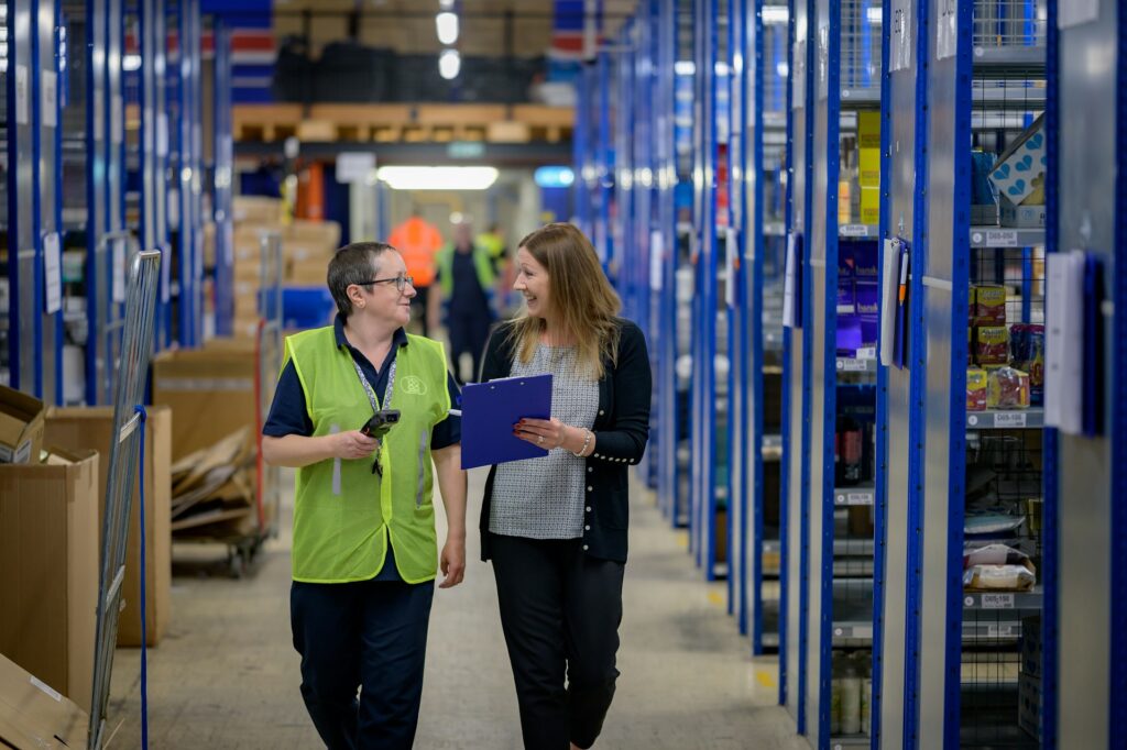 Two women walking through a warehouse and smiling, one dressed in a safety vest holding a scanner and the other in business attire while she holds a clipboard, discussing the impact of Sage Intacct on distribution operations