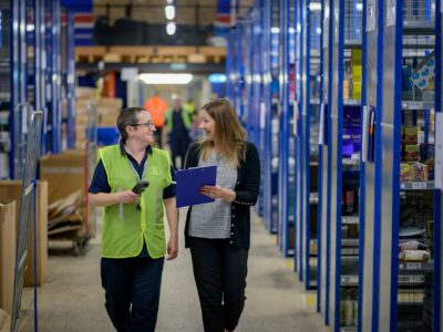 Two women walking through a warehouse and smiling, one dressed in a safety vest holding a scanner and the other in business attire while she holds a clipboard, discussing the impact of Sage Intacct on distribution operations