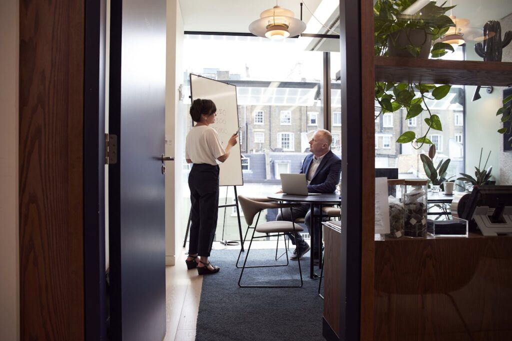 A zoomed out shot of an office with a man sitting and listening to a woman speak, who is a software consultant advising the man's ERP implementation.