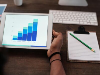 A hand holding a tablet displaying upwards fast growth January to May over a brown desk next to a notepad with a green pencil