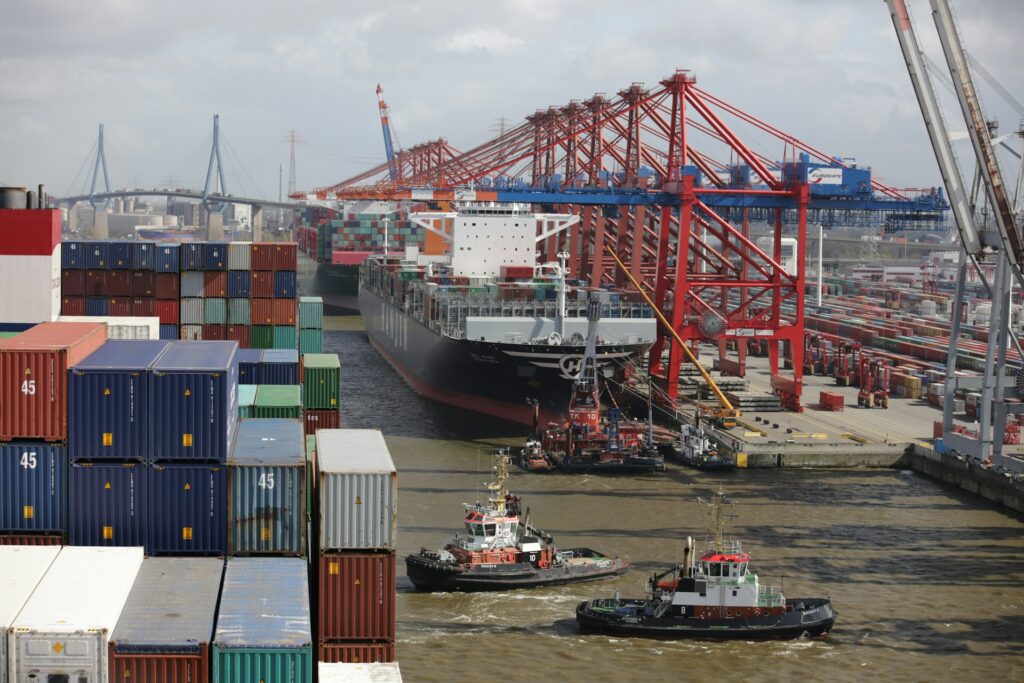 A port harbor with a docked ship, two tugboats, several loading cranes and stacks of crates representing the global supply chain  and visibiltiy achievable through Sage Intacct