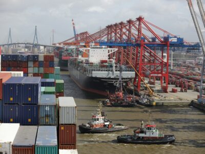 A port harbor with a docked ship, two tugboats, several loading cranes and stacks of crates representing the global supply chain and visibiltiy achievable through Sage Intacct