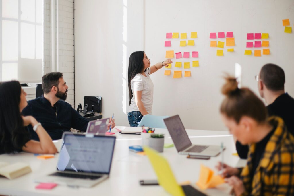 Officer workers review project management strategies using colorful sticky notes on a wall, with a woman in white pointing at a white board while four team members follow along on their laptops and discuss how ERP such as Acumatica or Deltek may help facilitate better project accounting and management for these use cases.