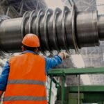 A factory worker in an orange safety vest and hard hat inspects a large industrial turbine shaft in a manufacturing facility, highlighting the type of operations that manufacturing ERP and accounting software helps optimize and monitor.