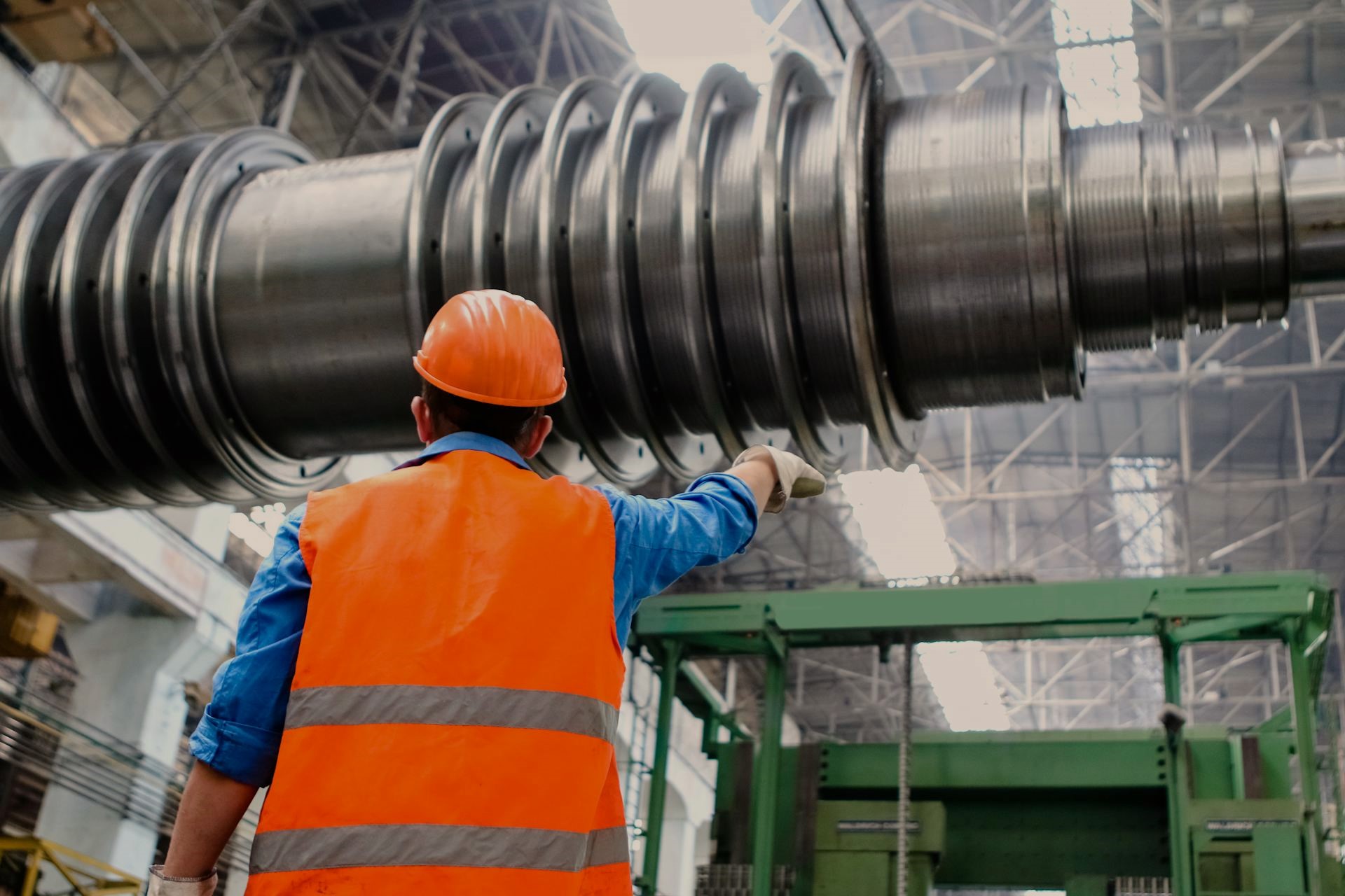 A factory worker in an orange safety vest and hard hat inspects a large industrial turbine shaft in a manufacturing facility, highlighting the type of operations that manufacturing ERP and accounting software helps optimize and monitor.