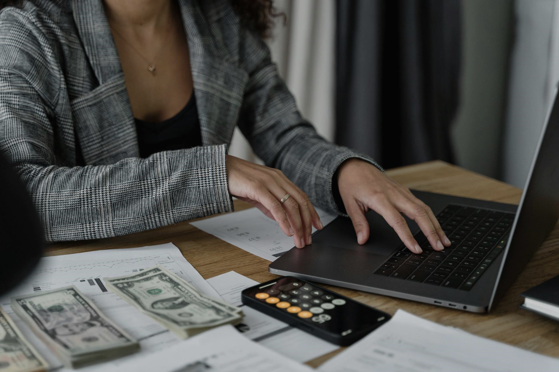 A professional in a plaid blazer works on AP automation software like Ottimate, with cash, financial documents, and a calculator spread across their wooden desk.