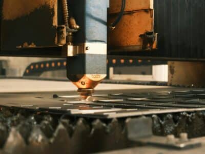 A close-up view of a laser cutting head in operation, showing the copper-colored nozzle cutting through metal with warm lighting highlighting the industrial machinery.