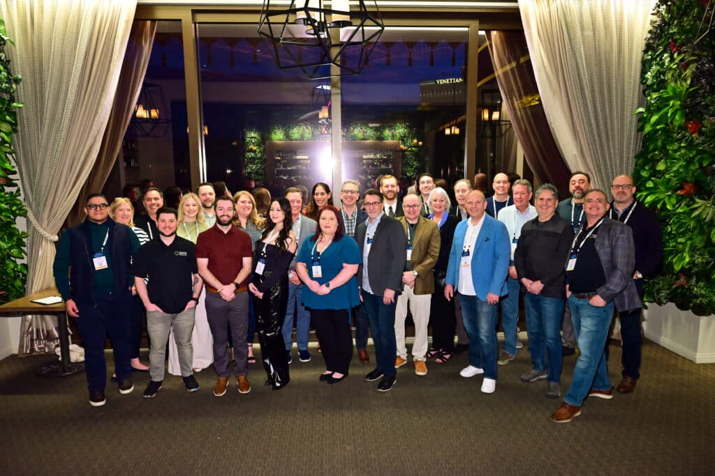 Group photo taken at La Cave in the Wynn Las Vegas during SWK Technologies' Customer Reception at Acumatica Summit 2025. Award recipients and SWK team members are gathered in front of an elegant venue entrance featuring white curtains and decorative greenery. Approximately twenty-five individuals are standing together in business casual attire, with the Venetian hotel visible through the windows in the background. 