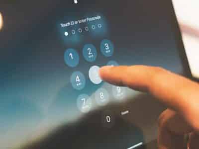 Person entering a passcode on a digital lock screen, touching the number 5 on a smartphone or tablet. The screen displays "Touch ID or Enter Passcode" with a numeric keypad, illustrating multi-factor authentication in action.