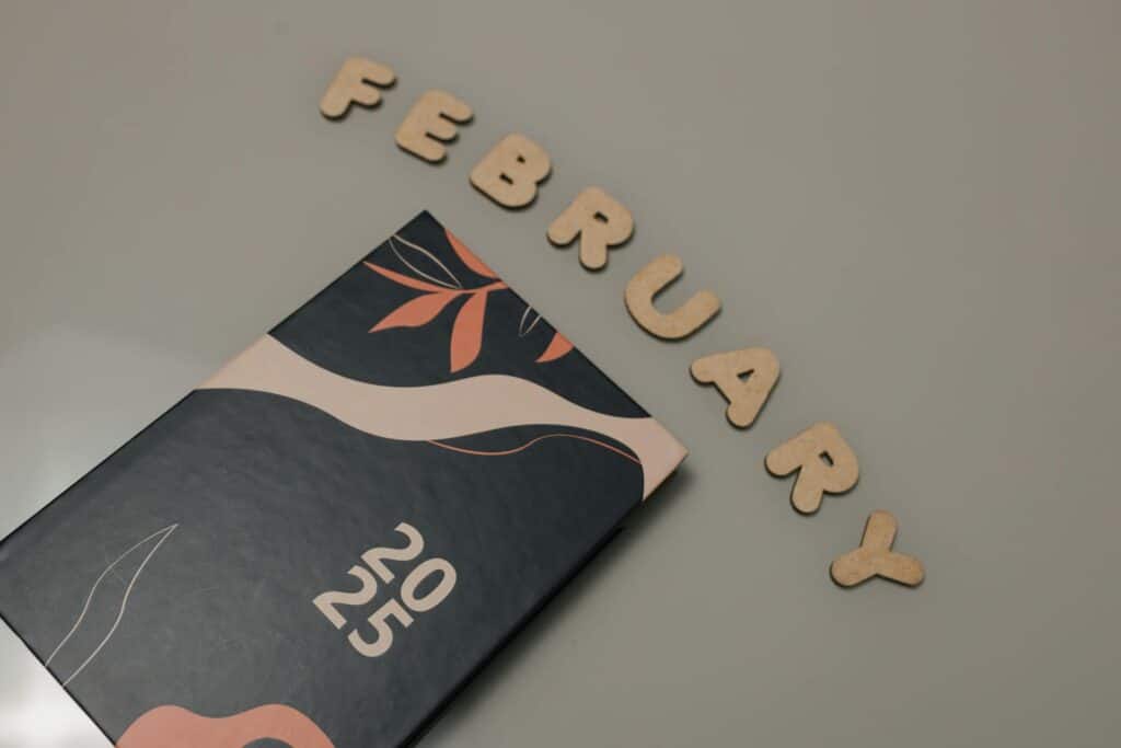 A stylized notebook or planner cover for 2025 in dark gray with beige and coral accents featuring abstract floral elements, with wooden letters spelling "FEBRUARY" arranged diagonally above it on a light gray surface.