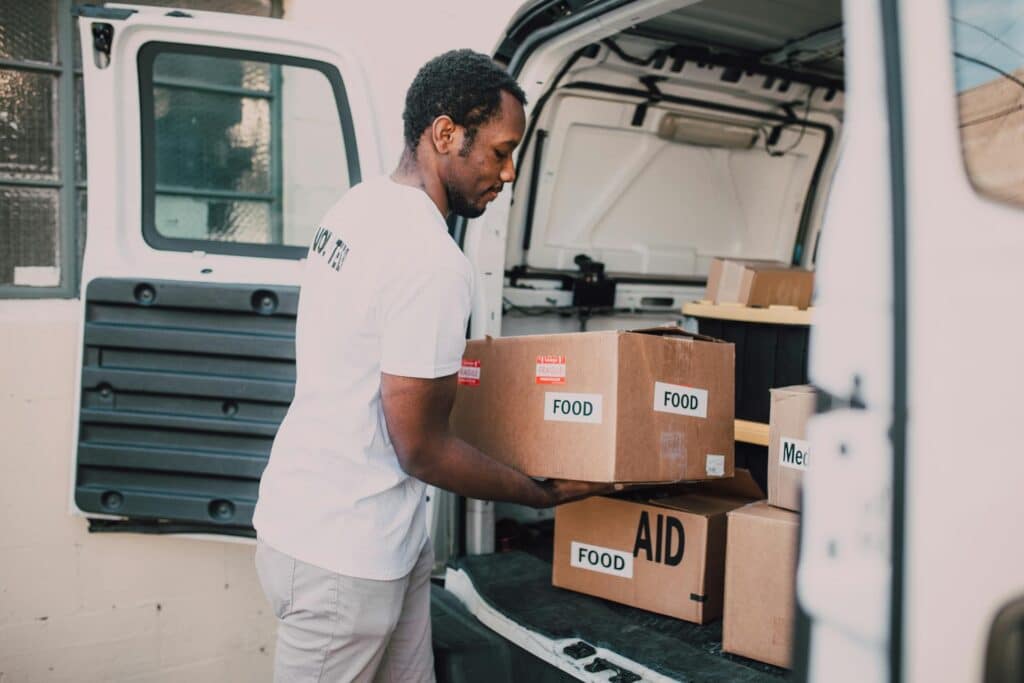 Nonprofit volunteer dressed in white t-shirt and grey pants loading boxes containing "food" and "aid" labels into the back area of a white van with doors open, representing the mission-critical activities that Sage Intacct's grant tracking capabilities help organizations monitor, manage and report on to ensure compliance and maximize impact.