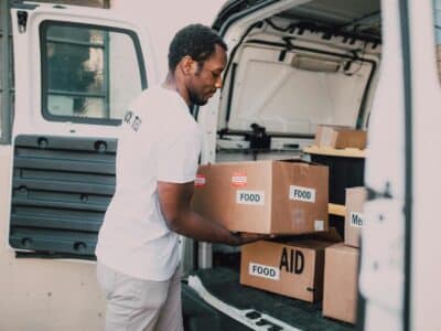 Nonprofit volunteer dressed in white t-shirt and grey pants loading boxes containing "food" and "aid" labels into the back area of a white van with doors open, representing the mission-critical activities that Sage Intacct's grant tracking capabilities help organizations monitor, manage and report on to ensure compliance and maximize impact.