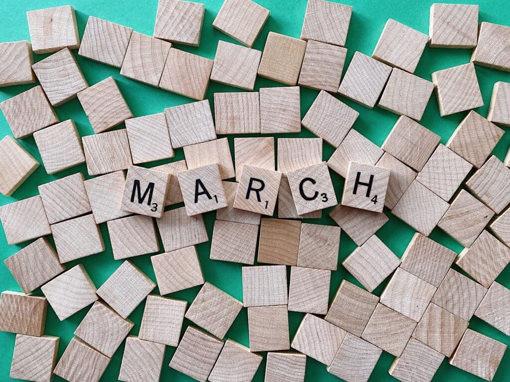 Wooden Scrabble tiles spelling "MARCH" arranged among numerous blank tiles on a bright teal background, representing SWK's March 2025 Cyber News Recap covering major security developments for the month.